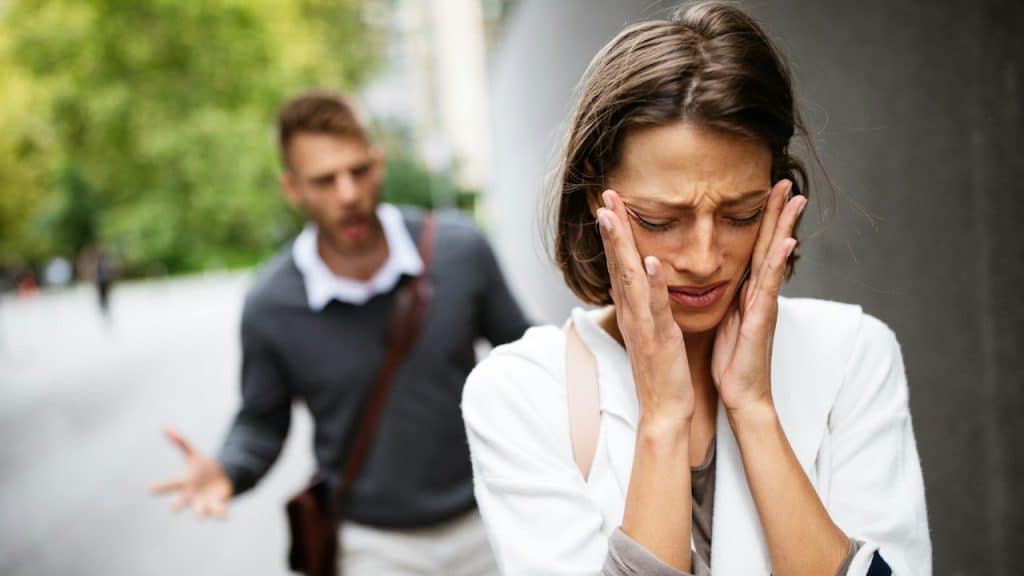 A woman crying and wiping her tears from her cheek with her hands as she stands with her back turned towards a man who’s gesturing at her.