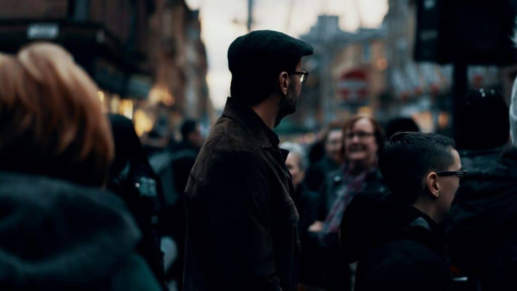 A man in a flat cap and glasses stands in a crowded, blurry city street at dusk.