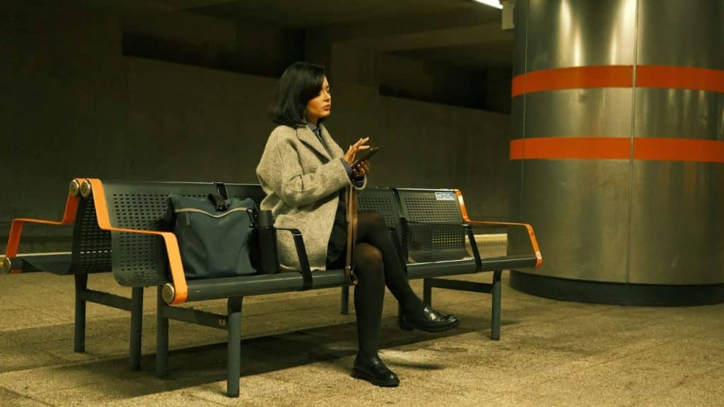 A woman in a grey coat sits on a subway bench while looking at her phone.