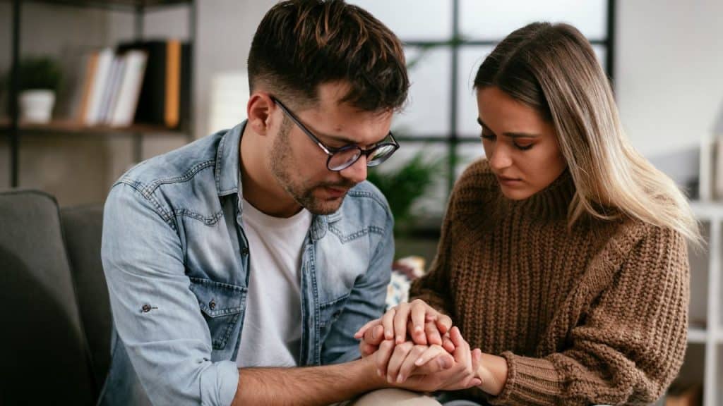 A man and woman sit close together indoors while holding both of each other's hands.