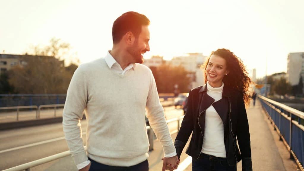 A man and woman hold hands and smile while walking on a bridge at sunset.