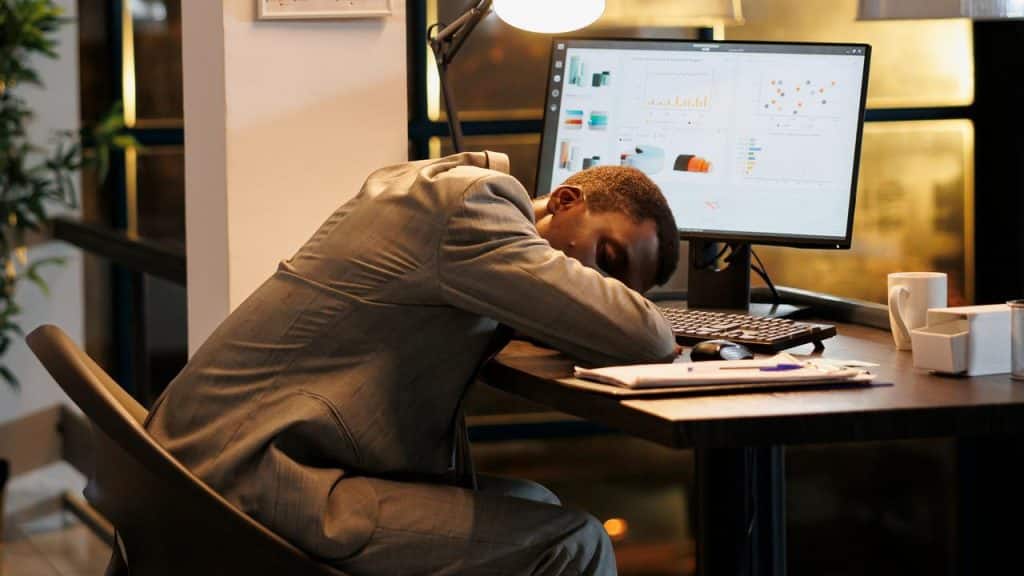 A tired office worker asleep at a desk in front of a computer monitor.