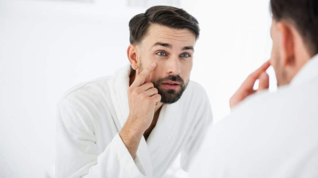 A man in a white robe stands before a mirror, touching the area below his eye.