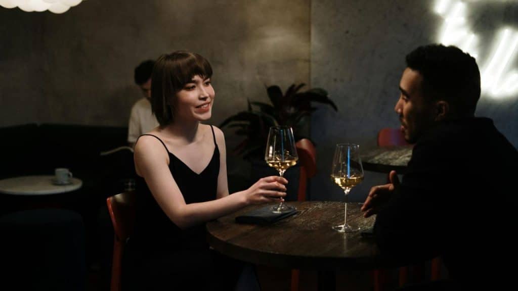 A woman in a black dress holds a wine glass while talking to a man..