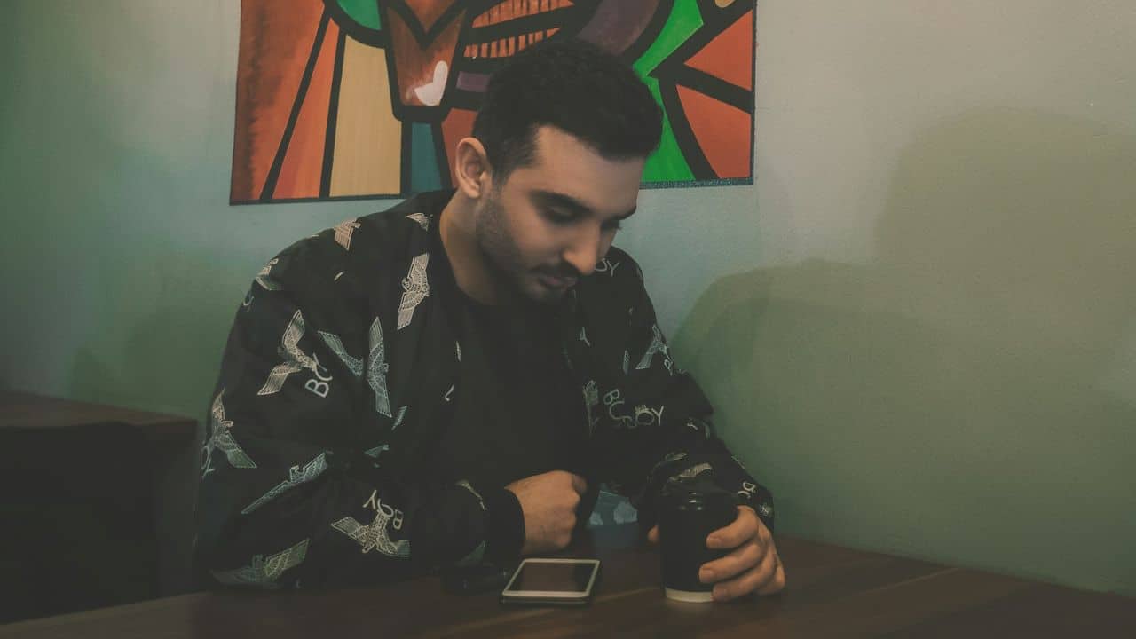 A man in a patterned jacket sits at a table looking down at a smartphone.
