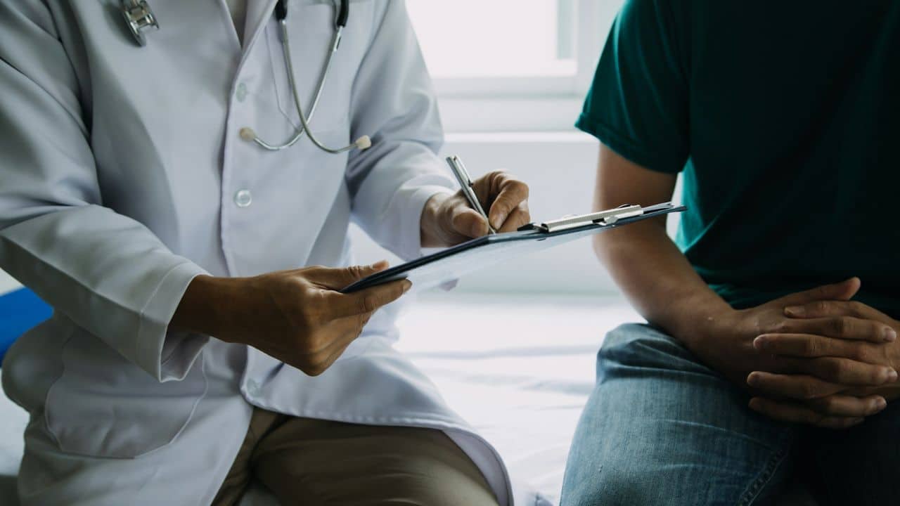 A doctor in a white lab coat writes on a clipboard while sitting with a patient.