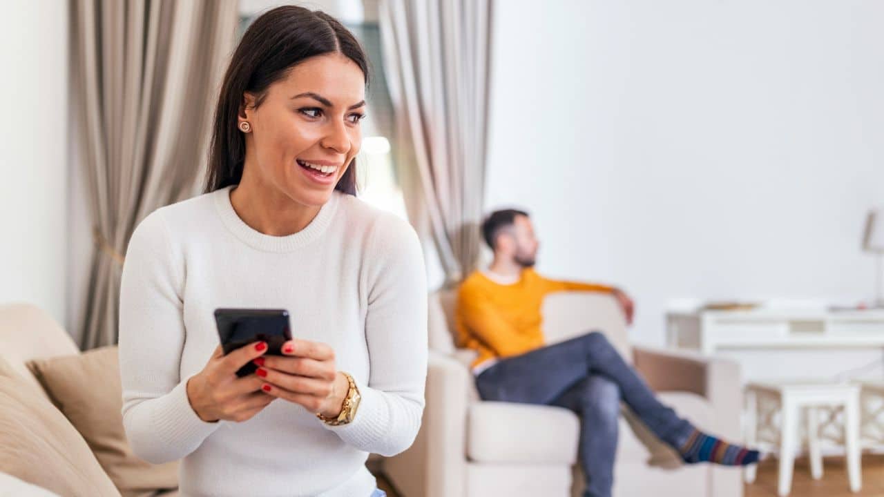 A woman smiles while holding a smartphone as a man sits in the background.