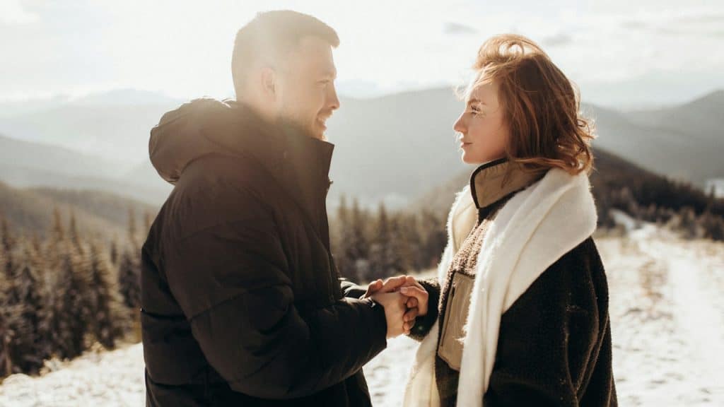 A man holding a woman’s hand while they stand on a snowy hill overlooking a forest.