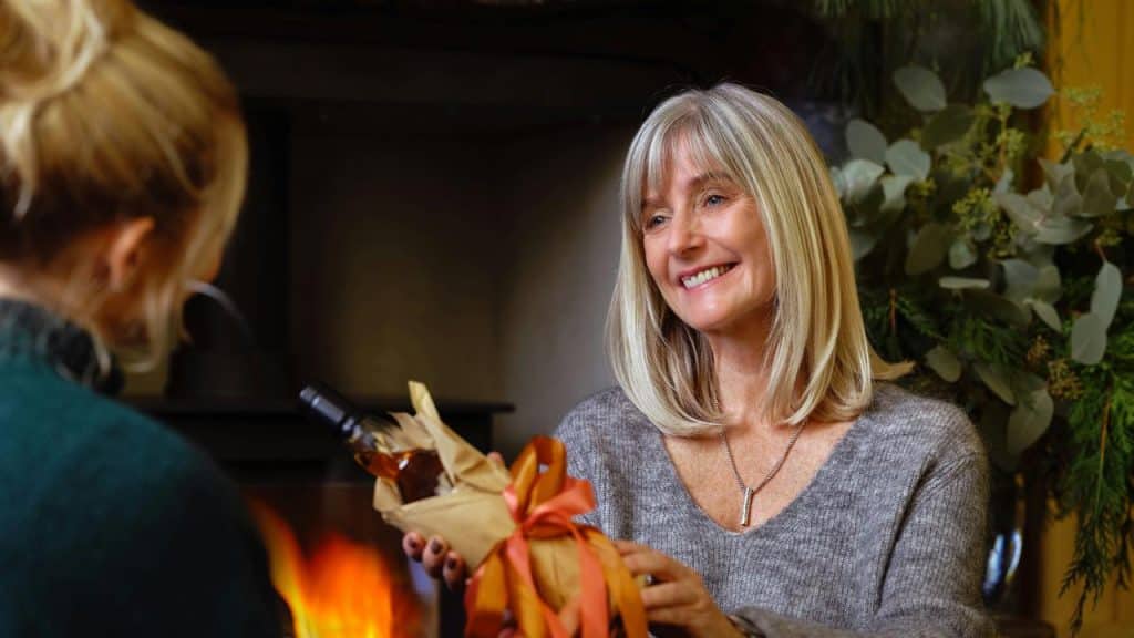A woman smiling while giving a wrapped gift to another person indoors.