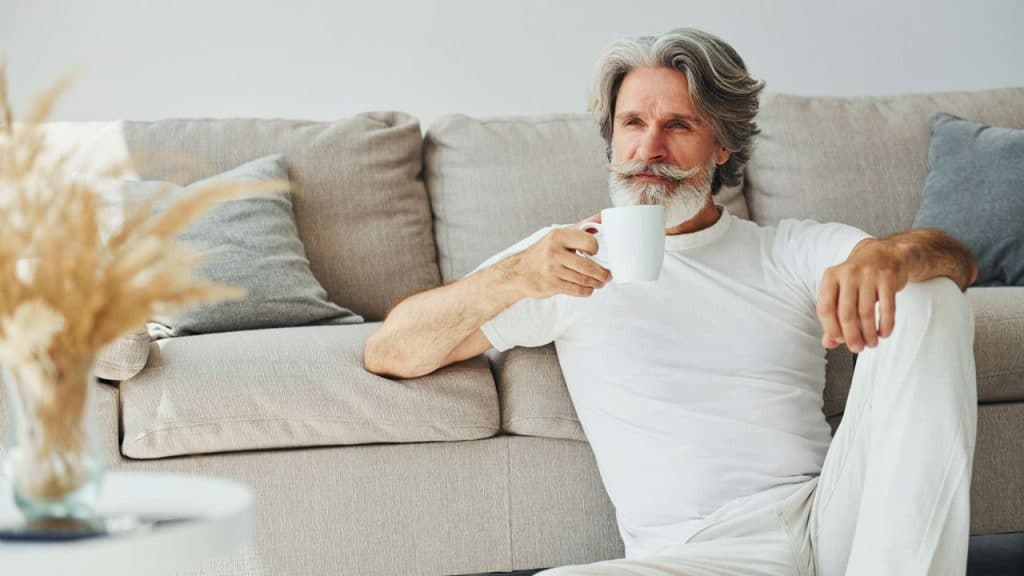 A grey-haired man with a mustache sits on the floor while holding a white mug.