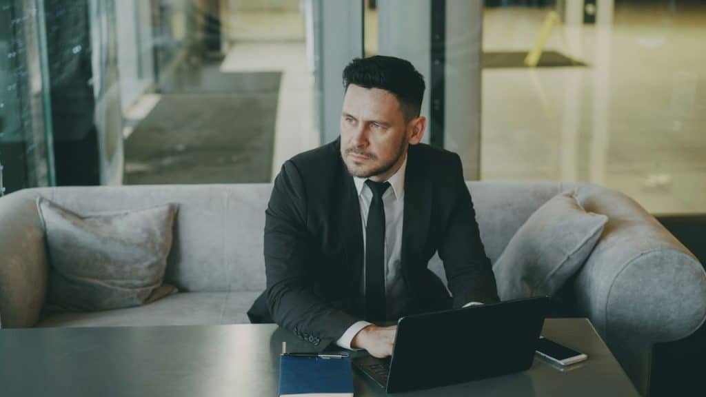 A man in a black suit and tie sits on a couch while using a laptop.