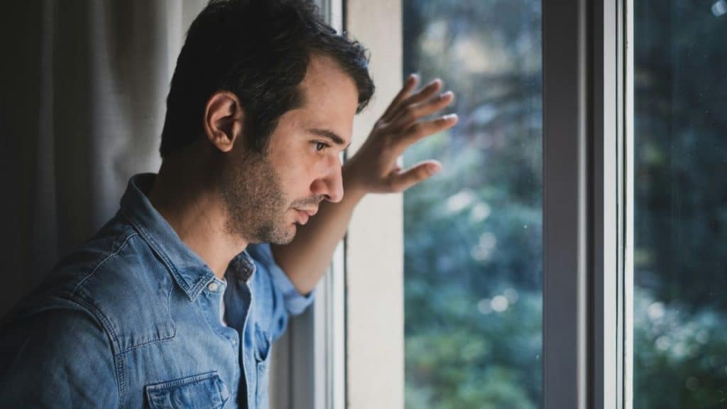 A man in a denim shirt looks out a window with his hand on the frame.