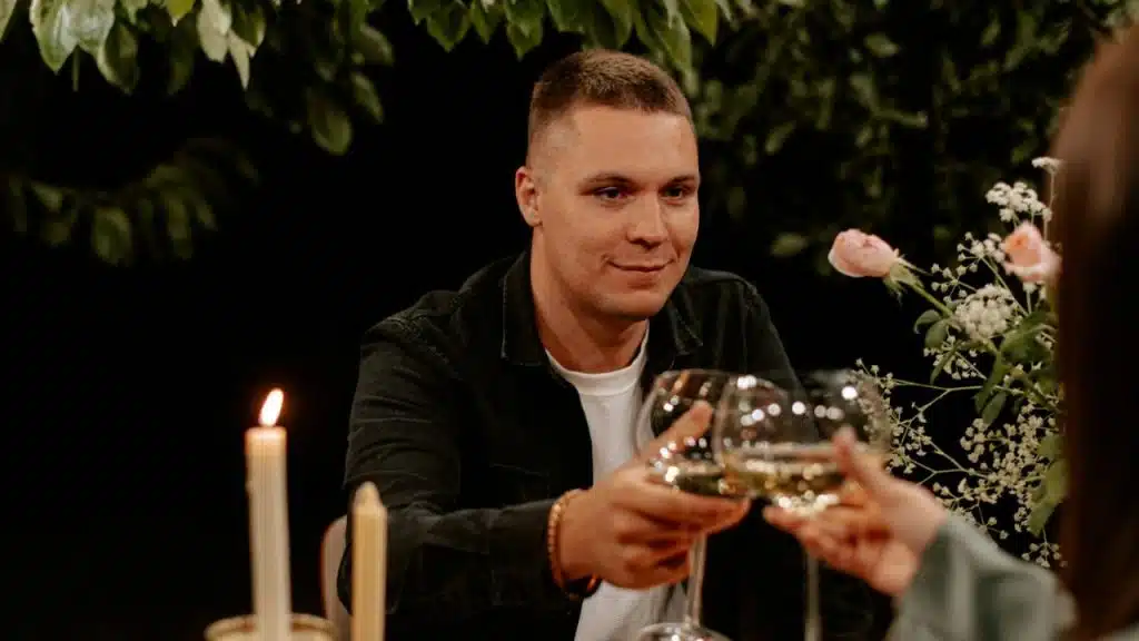 A man in a black shirt raises a wine glass to toast with a person off-camera.