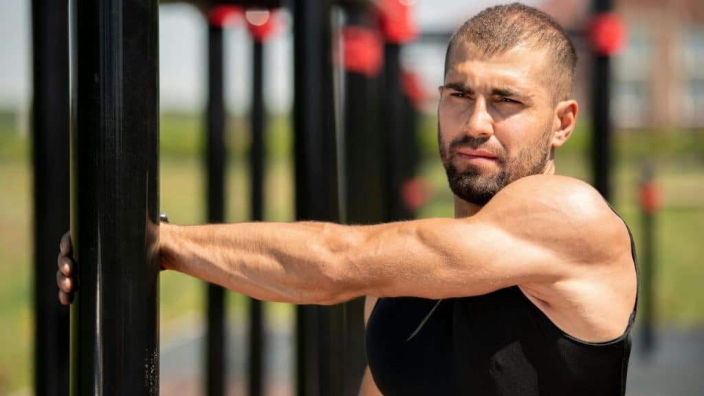 A muscular man with a beard and short hair stretches his arm at an outdoor gym.