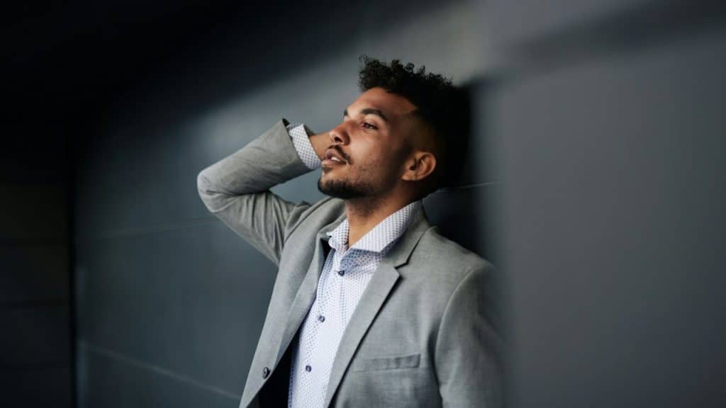 A man in a gray suit leans against a dark wall with his hand behind his head.