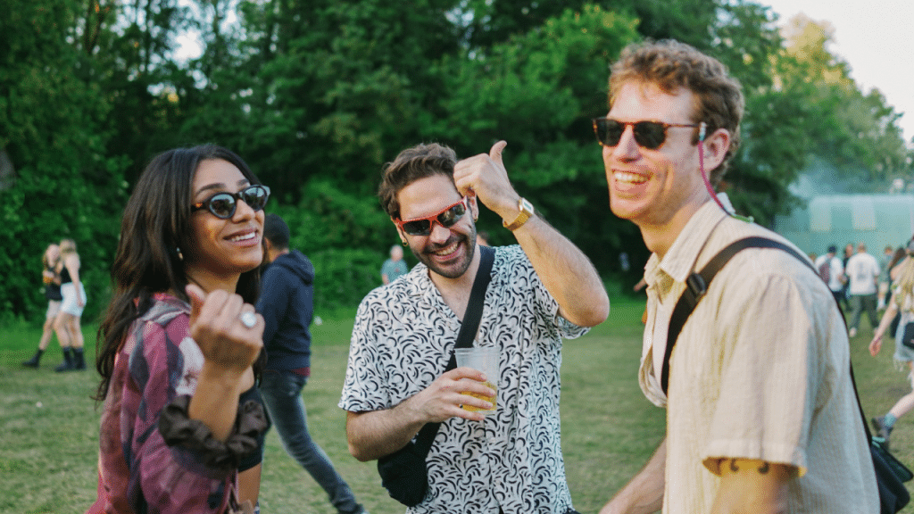 Three people wearing sunglasses smile and talk together in an open grassy outdoor field area.