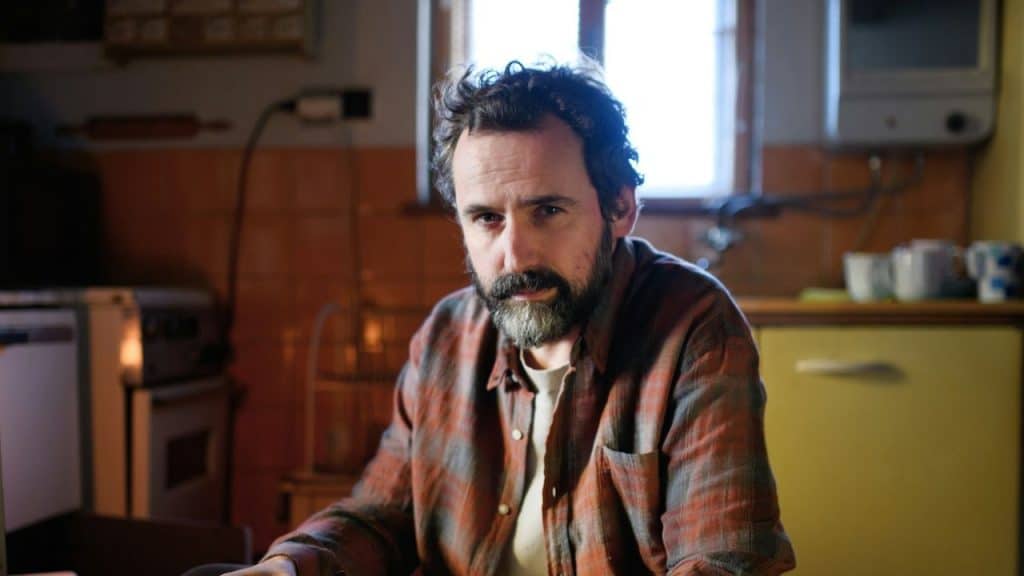 A man with a beard sits in a dimly lit kitchen looking directly forward.