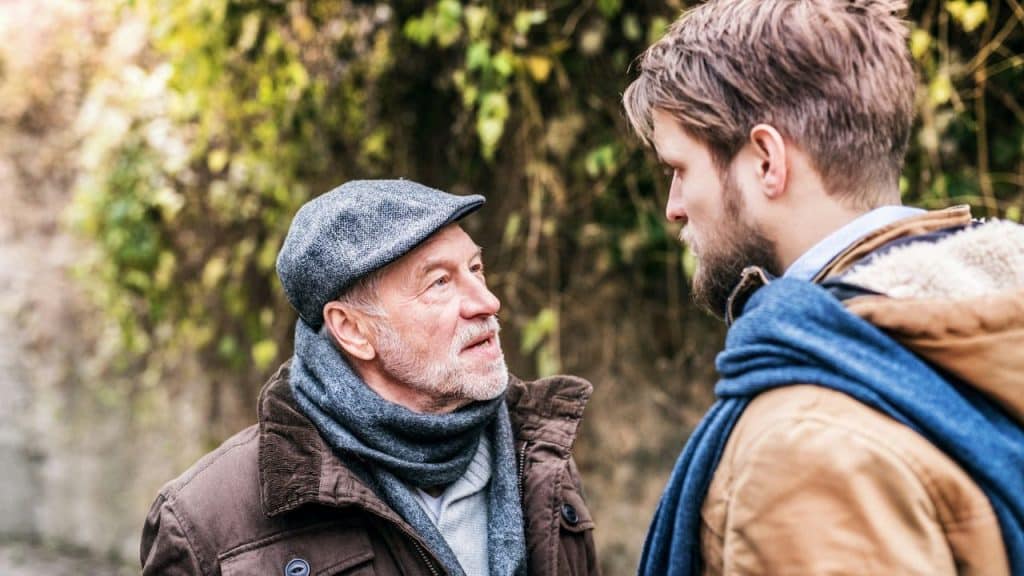 An older man in a flat cap and scarf looks up while talking to a younger man.