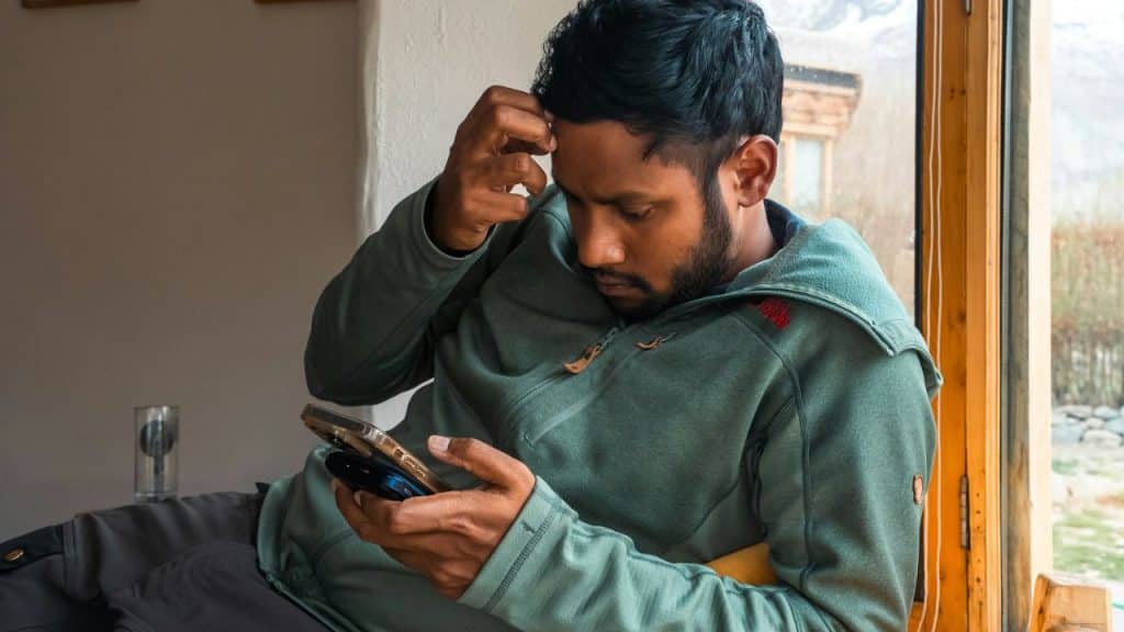 A man in a green zip-up jacket looks down at his phone while scratching his forehead.