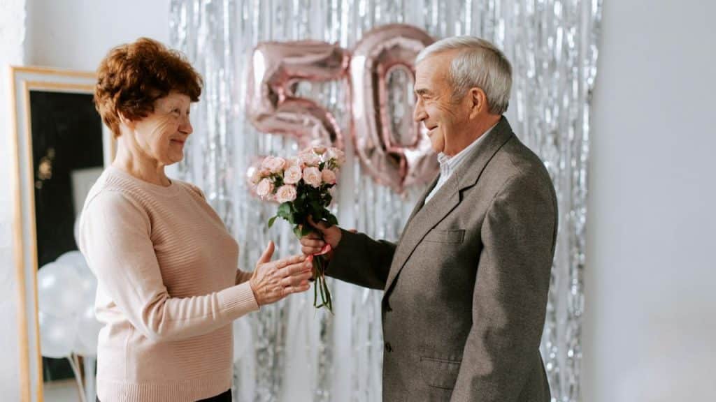 An elderly man in a suit gives pink roses to an elderly woman indoors.
