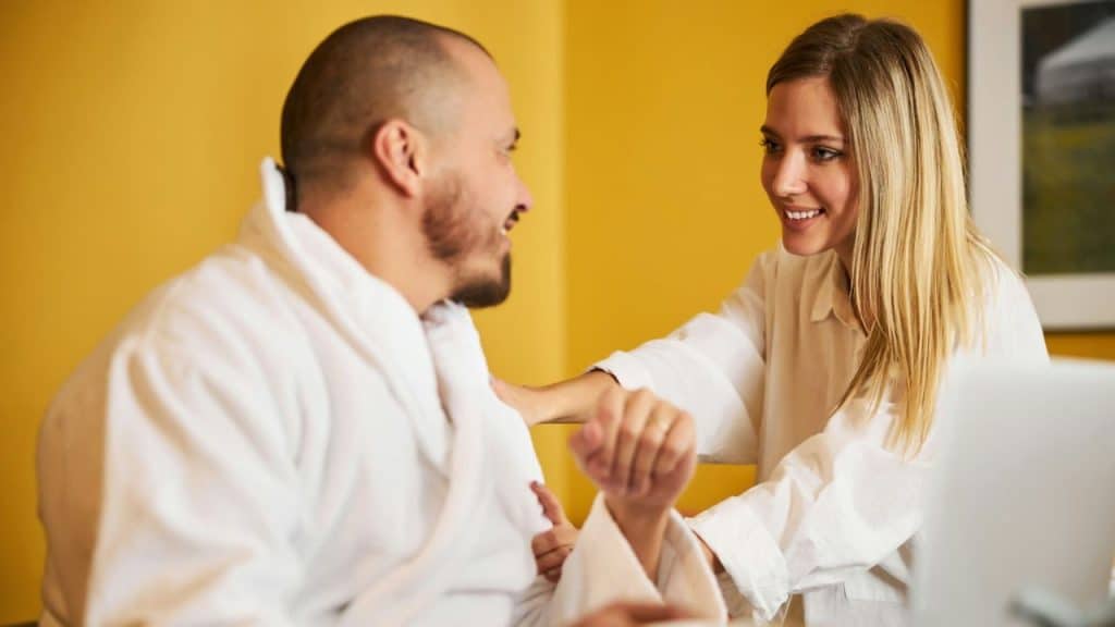 A woman in a white shirt smiles while touching the shoulder of a man in a bathrobe.