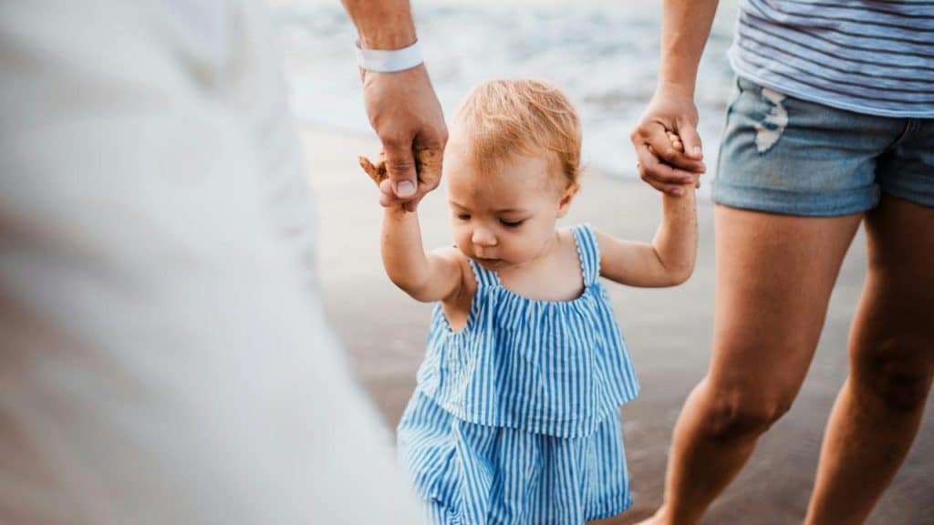 A toddler holding hands with two adults while walking.