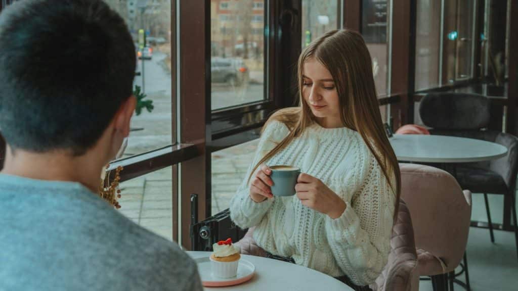 A woman in a white sweater holds a mug while sitting across from a man.
