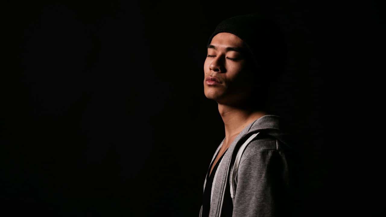A man in a beanie and hoodie stands with his eyes closed against a black background.