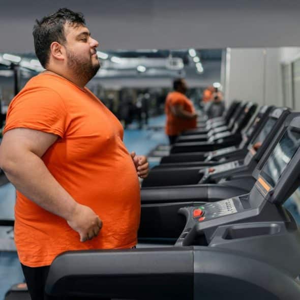 A man in an orange t-shirt walks on a treadmill in a modern indoor gym.
