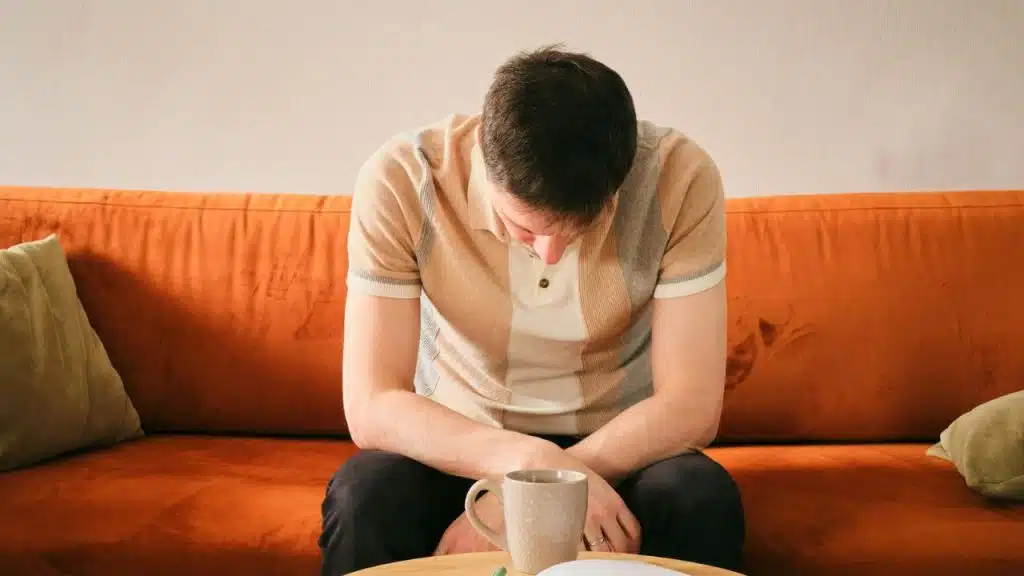 A man in a grey coat lies face down on an orange velvet sofa with a tired expression.