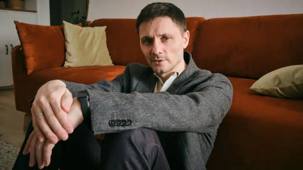 A man in a grey blazer sits on the floor in front of an orange sofa.