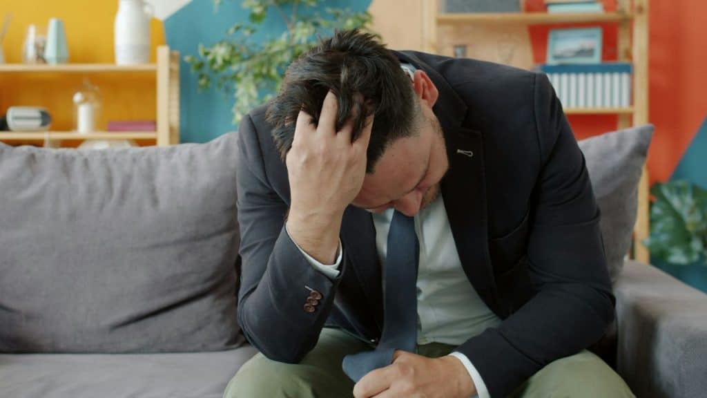 A man in a suit sits on a sofa, head in his hand.