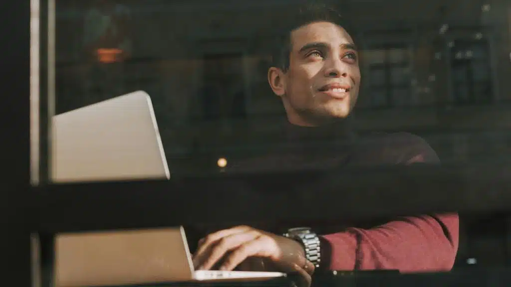 A man smiling while working on a laptop and looking out a window.