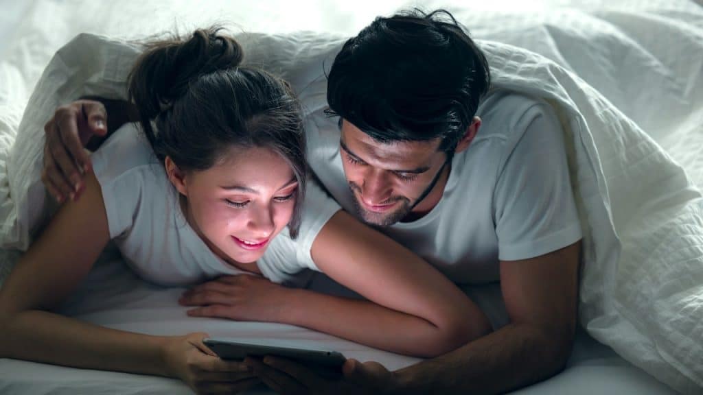 A young, sweet couple on a bed watching something on a tablet gadget and relaxing in the bedroom.