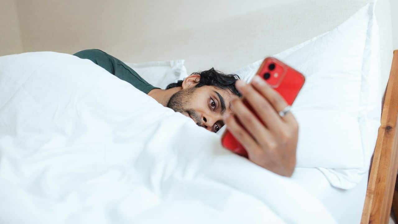 A man lies in bed under white covers, looking at a red smartphone in his hand.