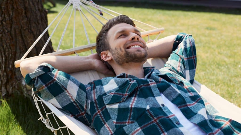 A bearded man resting his head on his hands and relaxing on a hammock outdoors.