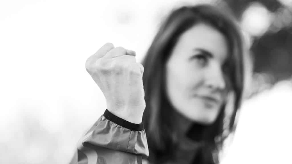 A black-and-white close-up of a person's clenched fist held up in front of their blurred face, symbolizing strength, determination, or resilience.