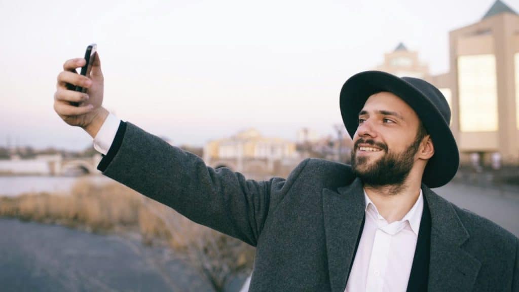 A bearded man in a hat and grey coat takes a selfie while smiling outdoors.