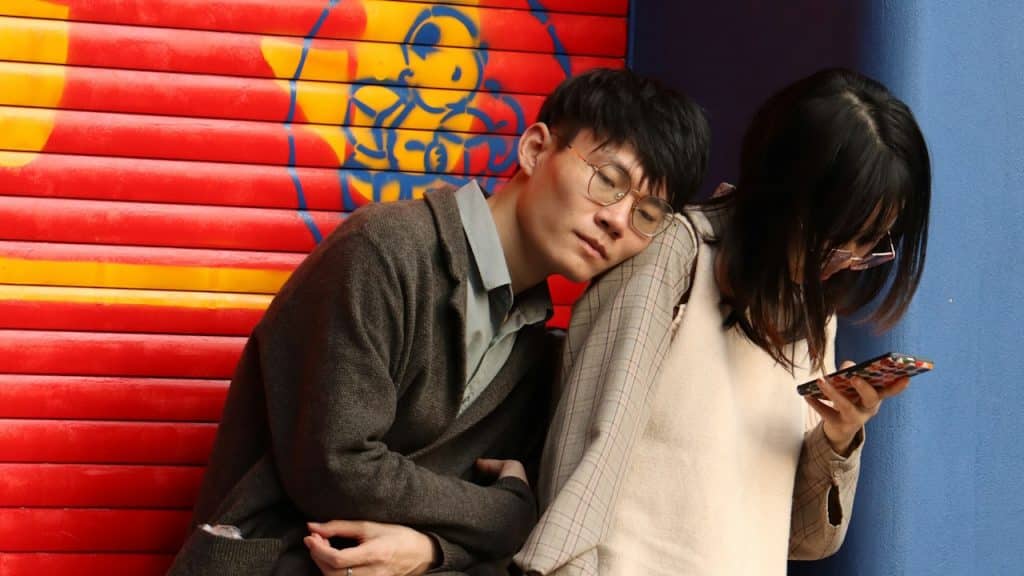 A man wearing glasses leaning on a woman’s shoulder while she scrolls through her phone.