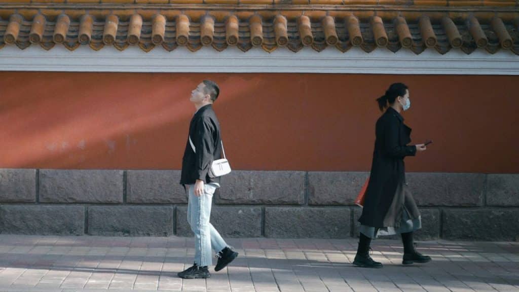 Two people walk in opposite directions on a sidewalk past a long orange and stone wall.