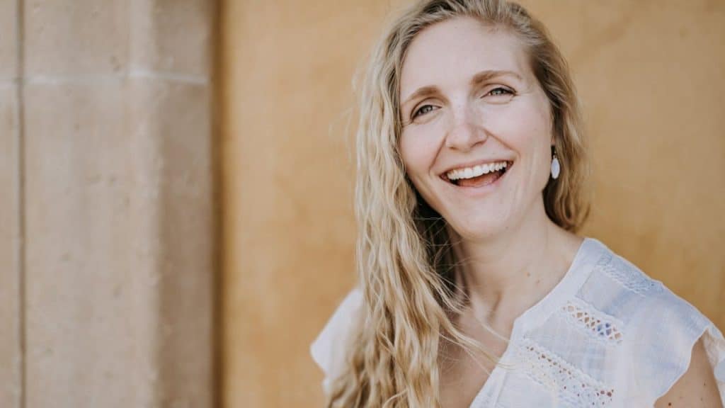 A laughing woman with long, wavy blonde hair stands in front of a textured yellow wall.