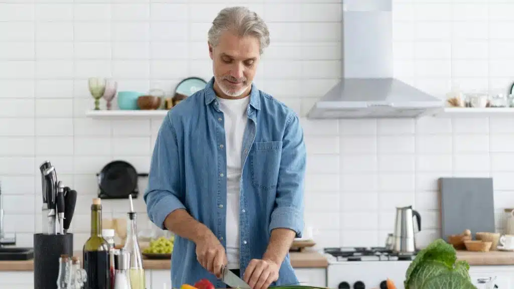 A handsome, mature man in a denim shirt is chopping vegetables in a clean, modern kitchen.