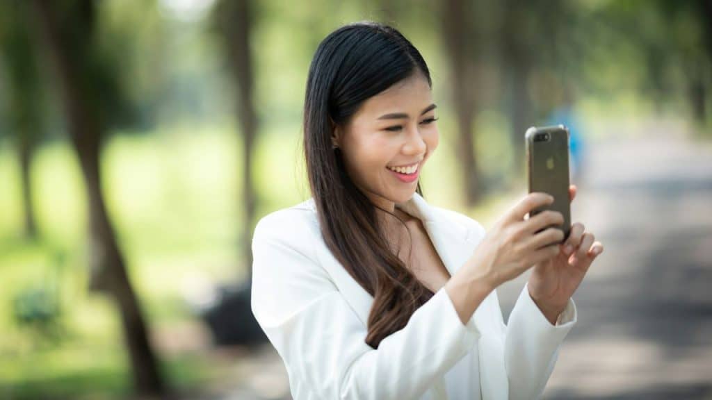 A smiling woman in a white blazer holds a smartphone in a park-like setting.