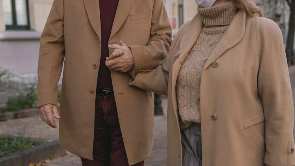 A man and a woman wearing matching tan overcoats hold hands while walking together on a brick-paved sidewalk.
