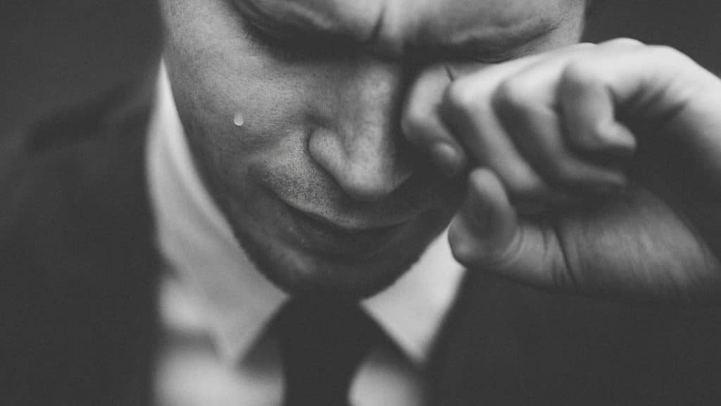 Close-up black and white photo of a man in a suit wiping a tear away.