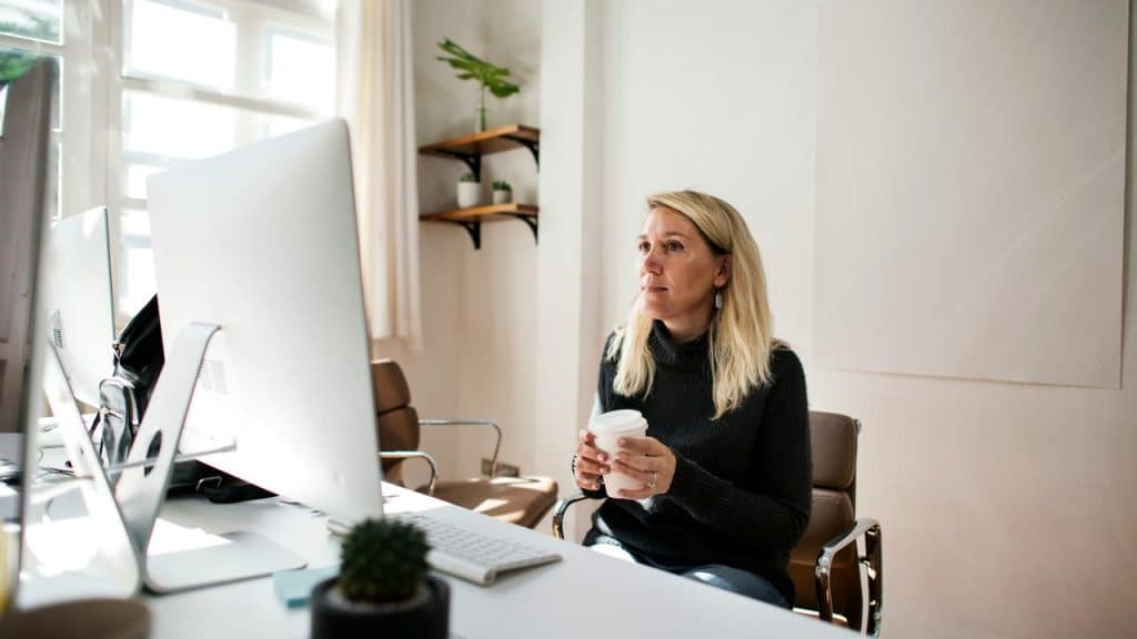 A woman sitting at a desk, holding a coffee cup and looking at a computer screen.