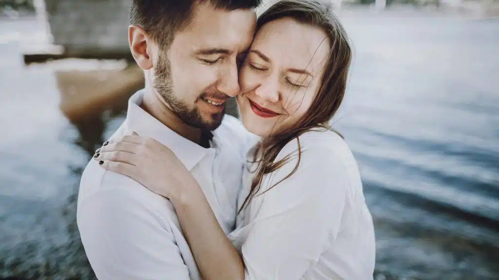 A couple in white shirts embracing closely by the water with their eyes closed.