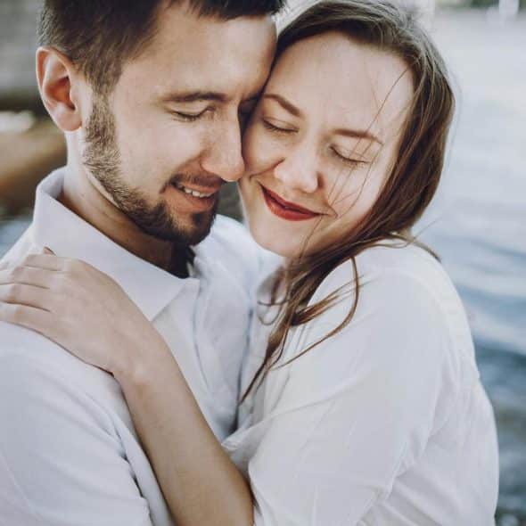 A couple in white shirts embracing closely by the water with their eyes closed.