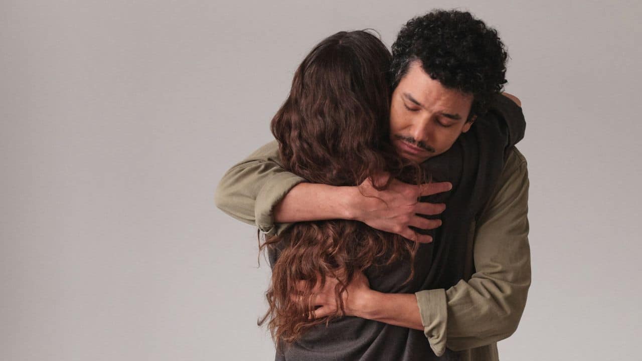 A man with curly hair and a mustache closes his eyes while hugging a woman.