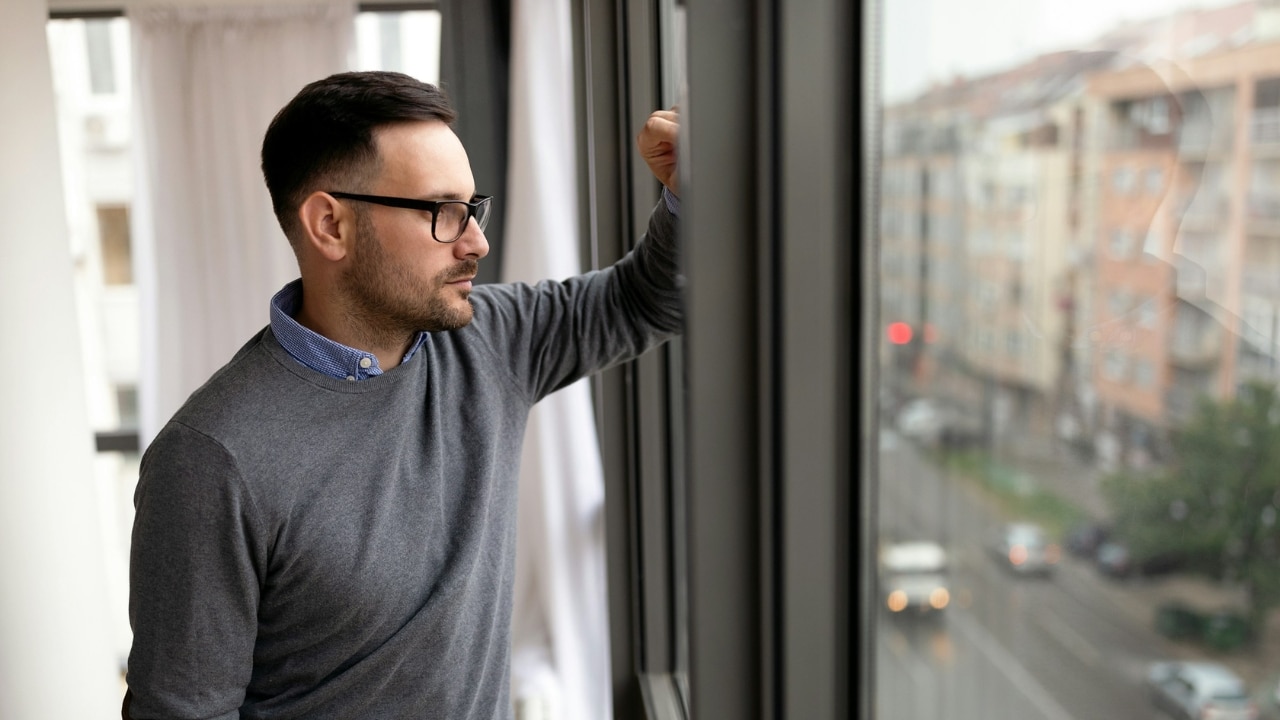 A man wearing glasses and a grey sweater looks out a window at a city.